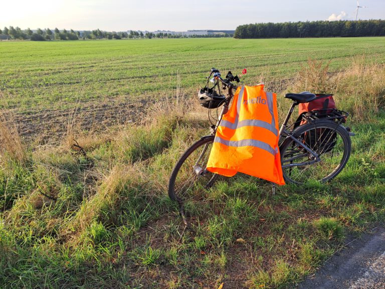 Zu sehen ist ein Fahrrad, das an einem sonnigen Tag an einem Feldrand steht. Am Lenker hängen Fahrradhelm und eine Warnweste in leuchtendem Orange. Am Gepäckträger ist eine Fahrradtasche befestigt.