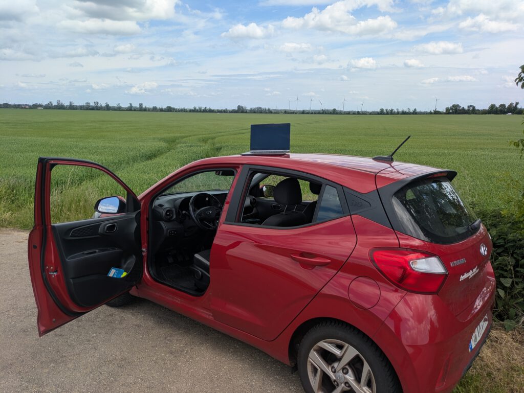 Ein rotes Auto vor einem Feld. Auf dem Dach steht ein aufgeklappter Laptop, der der Dokumentation der Hausbegehungen dient.