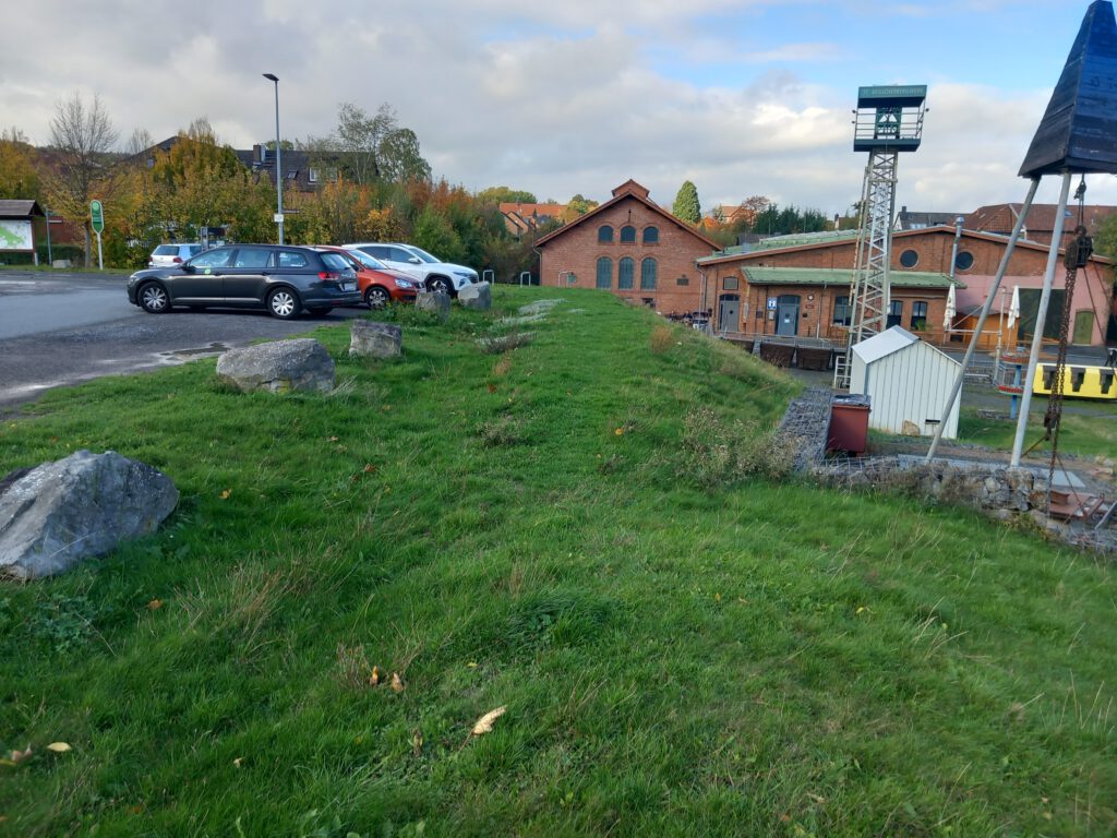 Das Bild zeigt einen Teil des Parkplatzes mit einzelnen Pkw vor der Umgestaltung. Im Hintergrund ist das Besucherberkwerk Klosterstollen zu sehen.