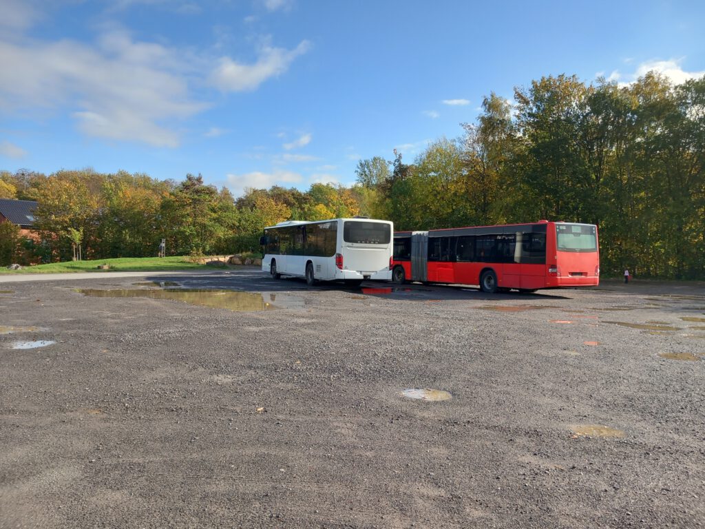 Das Bild zeigt den Parkplatz vor der Umgestaltung. Geparkt sind zwei Reisebusse auf einer Schotterfläche mit großen Regenpfützen
